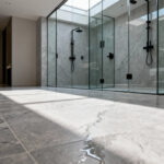 Seamless luxury bathroom flooring made of high-density porcelain tiles replicating the organic grain of honed taupe limestone in a walk-in shower setting.