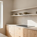 Bright, minimalist Scandinavian kitchen with light wood cabinetry, honed stone countertops, integrated induction hob, and open shelving featuring ceramic bowls. Natural light fills the serene, clutter-free space.