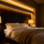 A photograph of a luxurious, modern primary bedroom bathed entirely in deep golden light (2800K warm color temperature), illustrating foundational lighting designed to promote rest and sleep.