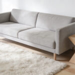 A wide-angle view of a minimalist living room showcasing ample negative space with white walls, a light gray linen sofa, and natural wood flooring, emphasizing organization and visual calmness to reduce cognitive load.